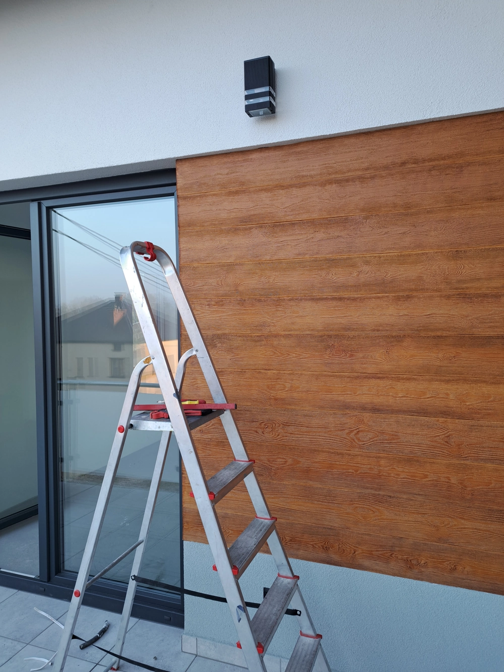 Drabina przed elewacją panelową z zamontowanym kinkietem LED podczas prac instalacji oświetlenia