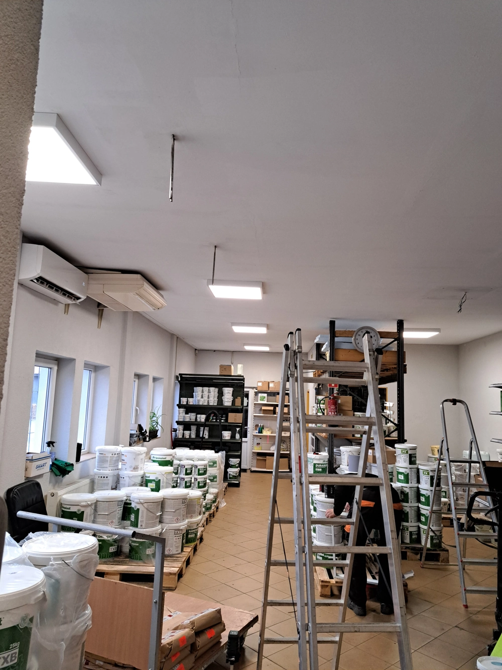 Wnętrze magazynu z regałami i pojemnikami, drabiną oraz zamontowanymi sufitowymi panelami LED – Oświetlenie